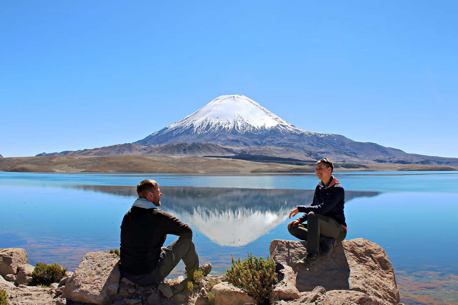 Johanna y Nils sentados frente al lago Chungará con el volcán Parinacota reflejado en el agua