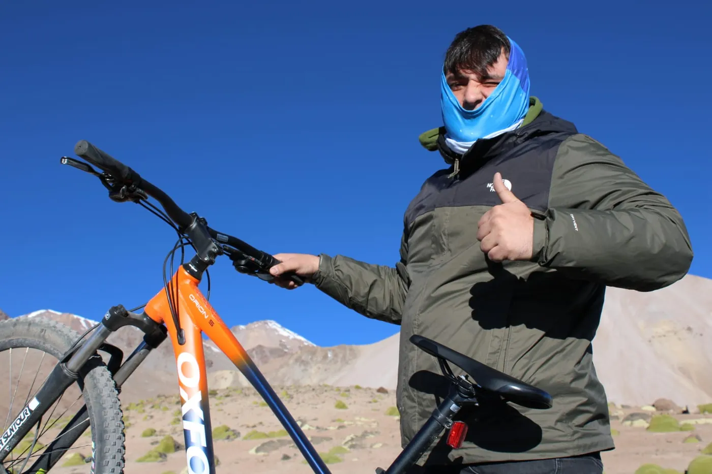 Ciclista con cubrebocas dando el pulgar arriba junto a su bicicleta de montaña, cordillera al fondo