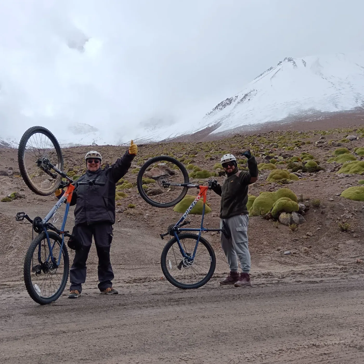 Ciclistas celebrando con las bicicletas en alto, volcán nevado entre nubes de fondo