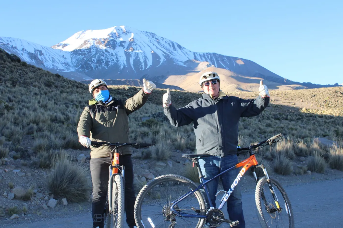 Dos ciclistas celebrando con los brazos en alto, volcán nevado entre nubes de fondo