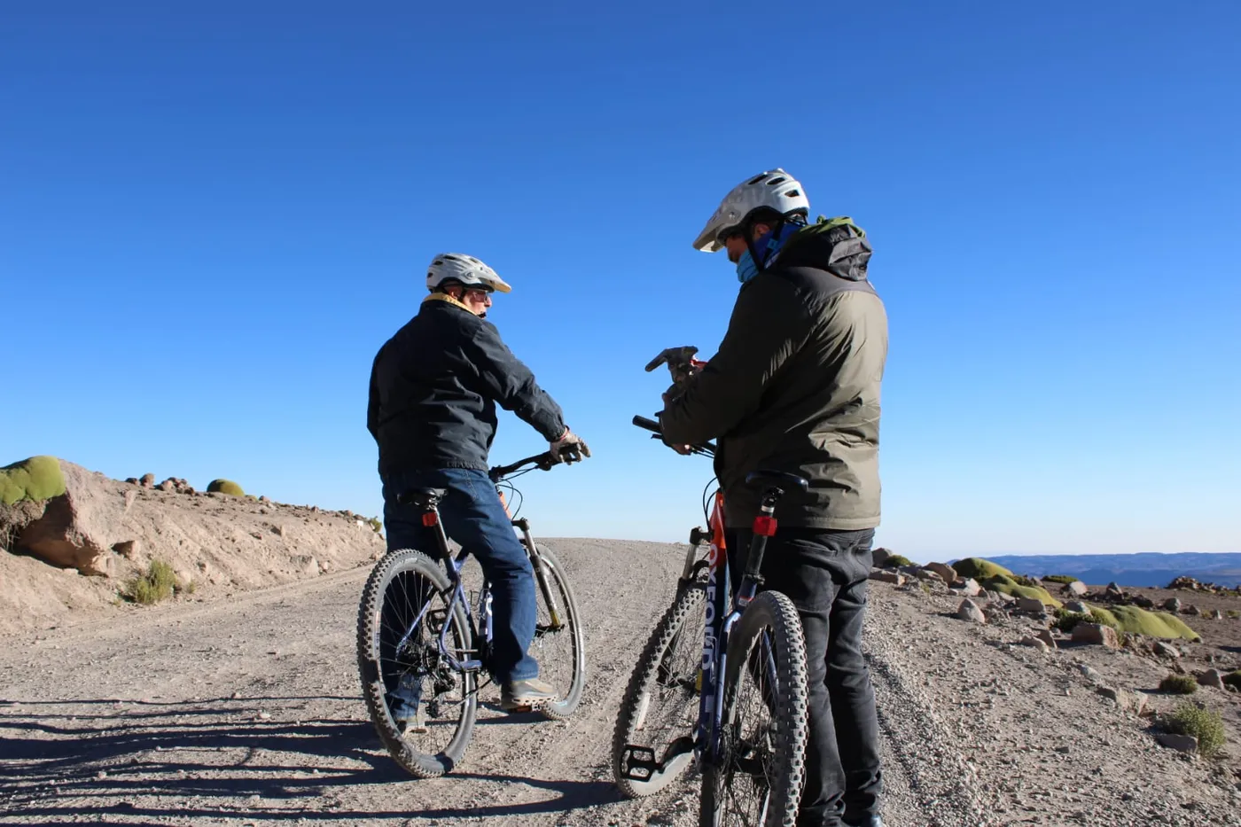 Dos ciclistas alejándose por camino de ripio al pie de una gran ladera montañosa